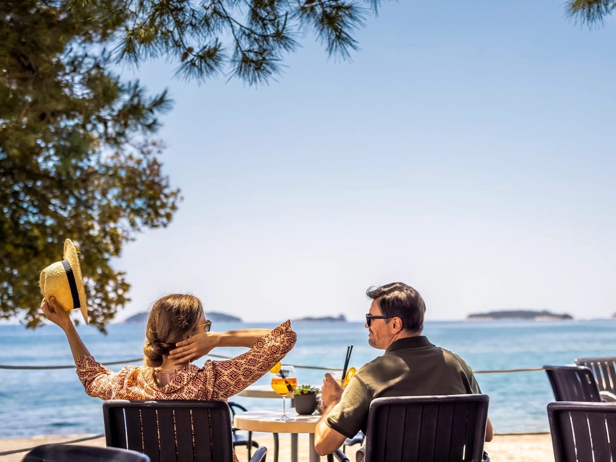 A couple with drinks at a table on a paved terrace beside calm blue sea, a pine bough overhead and islands on the horizon.