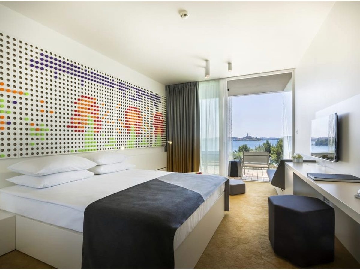 Hotel room with a coloured peg-board wall, a double bed, a desk and open glass doors to a balcony with sea and a distant old town.