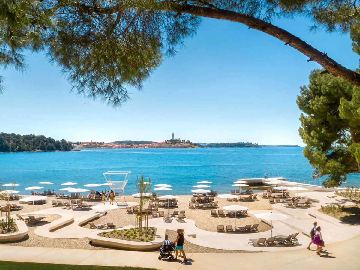 A landscaped pebble and concrete beach with cream parasols and loungers, calm blue sea and the terracotta roofs and spire of Rovinj across the bay, framed by a pine bough.