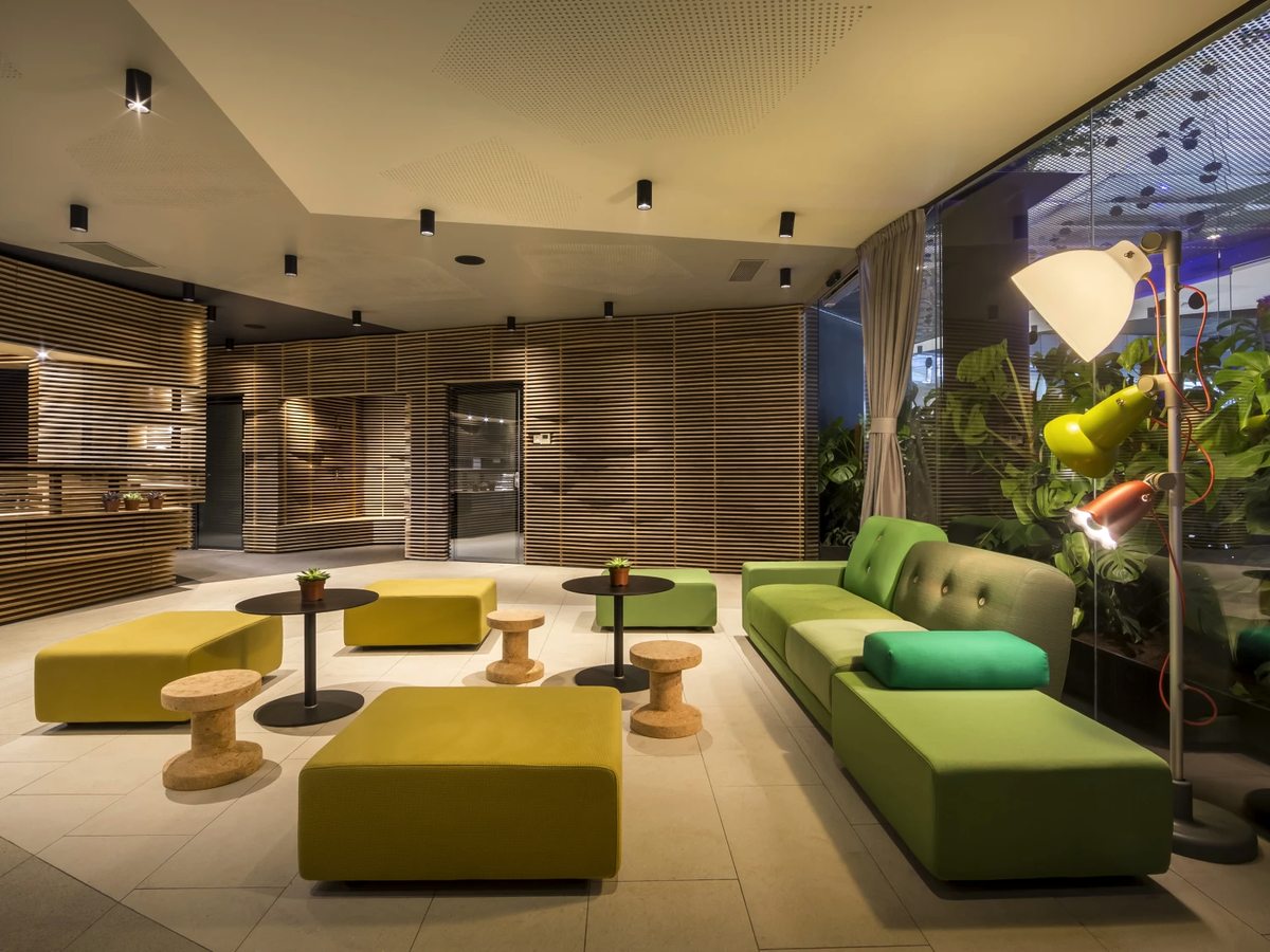 Lobby lounge with lime and green upholstered benches, cork stools, small round tables and timber-slatted walls, with plants behind glass.