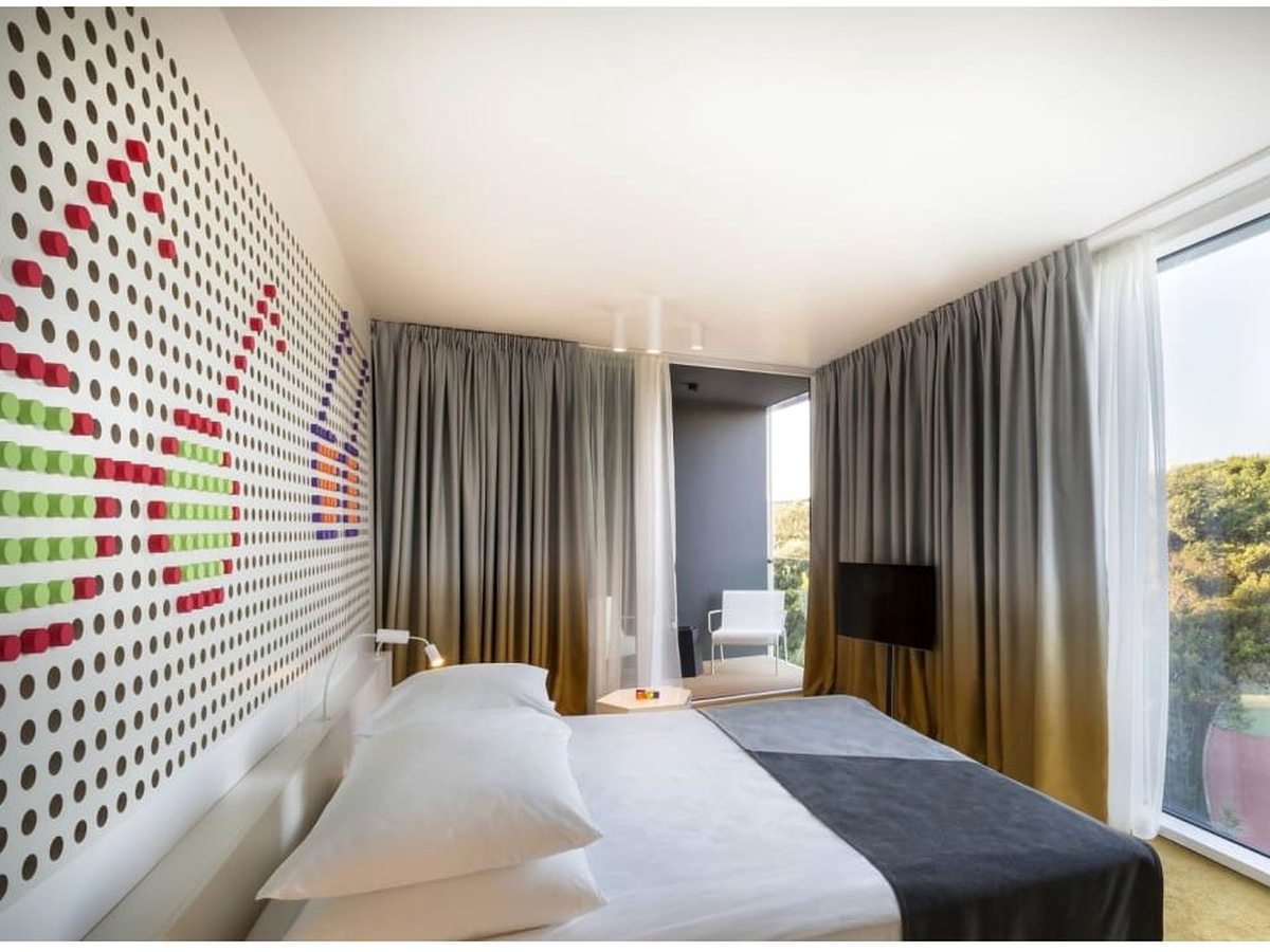 Hotel room with a peg-board headboard, a double bed and full-height windows onto a balcony backed by green woodland.