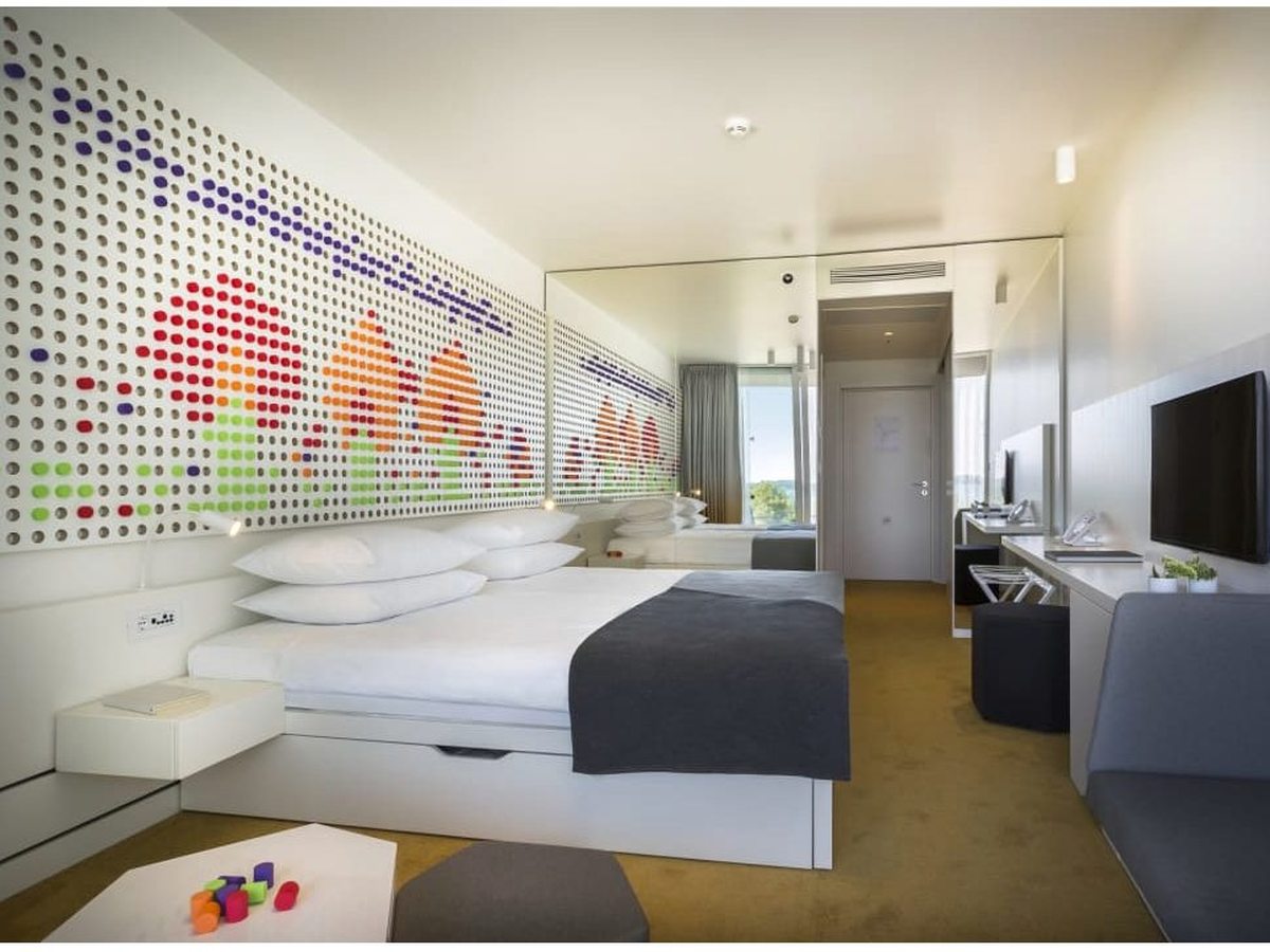 Hotel room with a white peg-board headboard patterned in red, orange, green and purple pegs, a double bed, a grey sofa and toys on the floor.