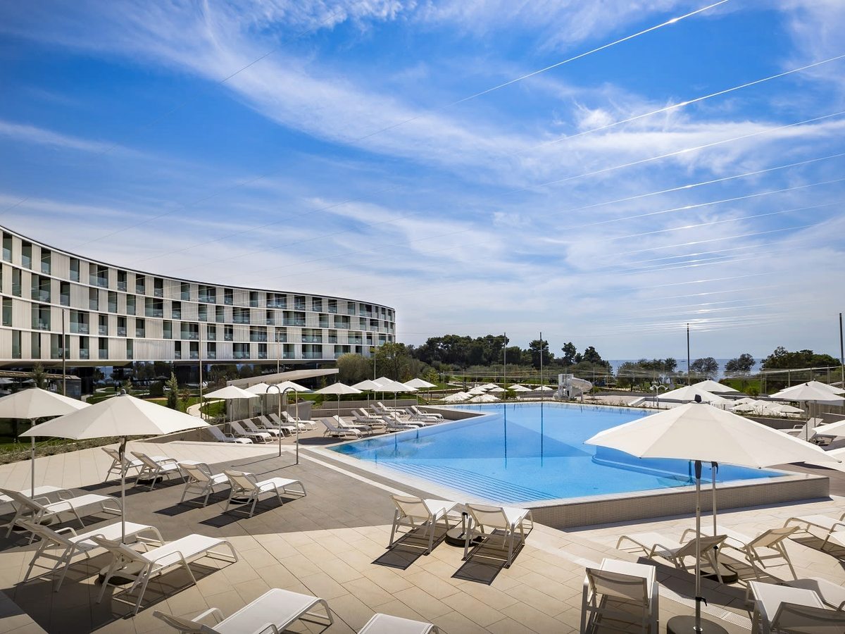 Rectangular outdoor pool with white parasols and rows of sun loungers on a pale stone deck, the curved white hotel behind under a blue sky.