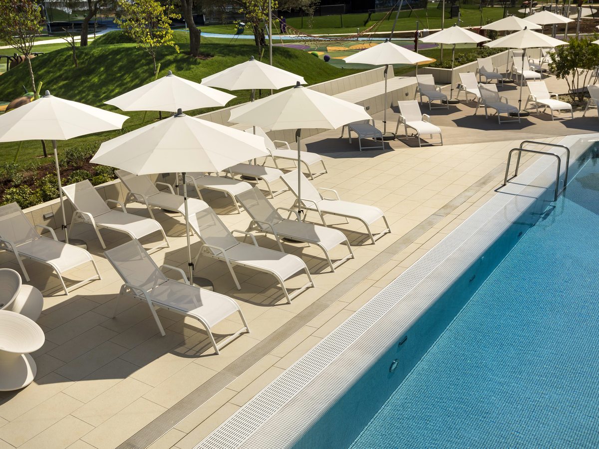 Rows of white sun loungers and cream parasols on the paved surround of a turquoise outdoor pool, with lawns and a playground behind.