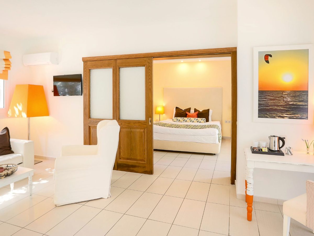 White-tiled suite living area with a sofa, armchair, wall-mounted television and a tea and coffee tray on a side table, with wooden sliding doors open to the bedroom beyond.