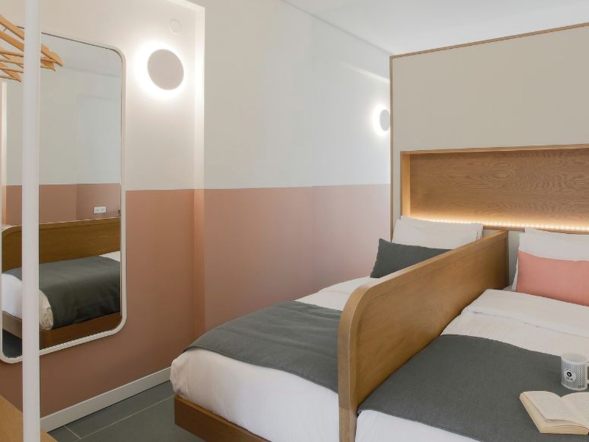 Compact bedroom with two single beds against a wooden backlit headboard, a pink and white two-tone wall, a full-length mirror and a hanging clothes rail.