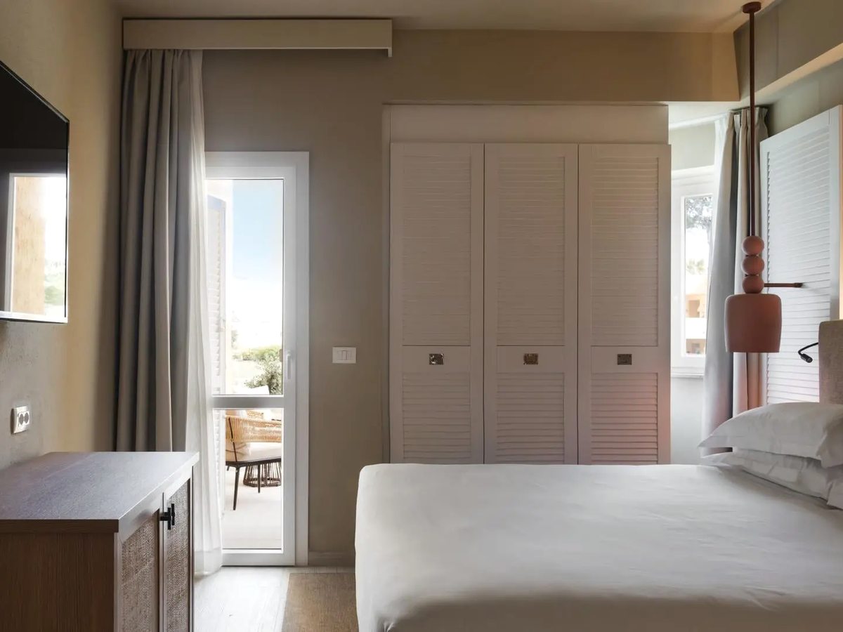 A double room with a wooden console and television, louvred wardrobe doors and a glazed door standing open onto a small balcony with a wicker chair.