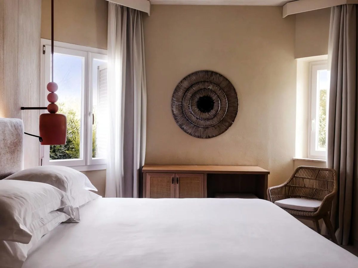 A double room with white bedding, a woven circular wall hanging above a low wooden sideboard, a wicker chair and two windows filled with sunlit garden foliage.