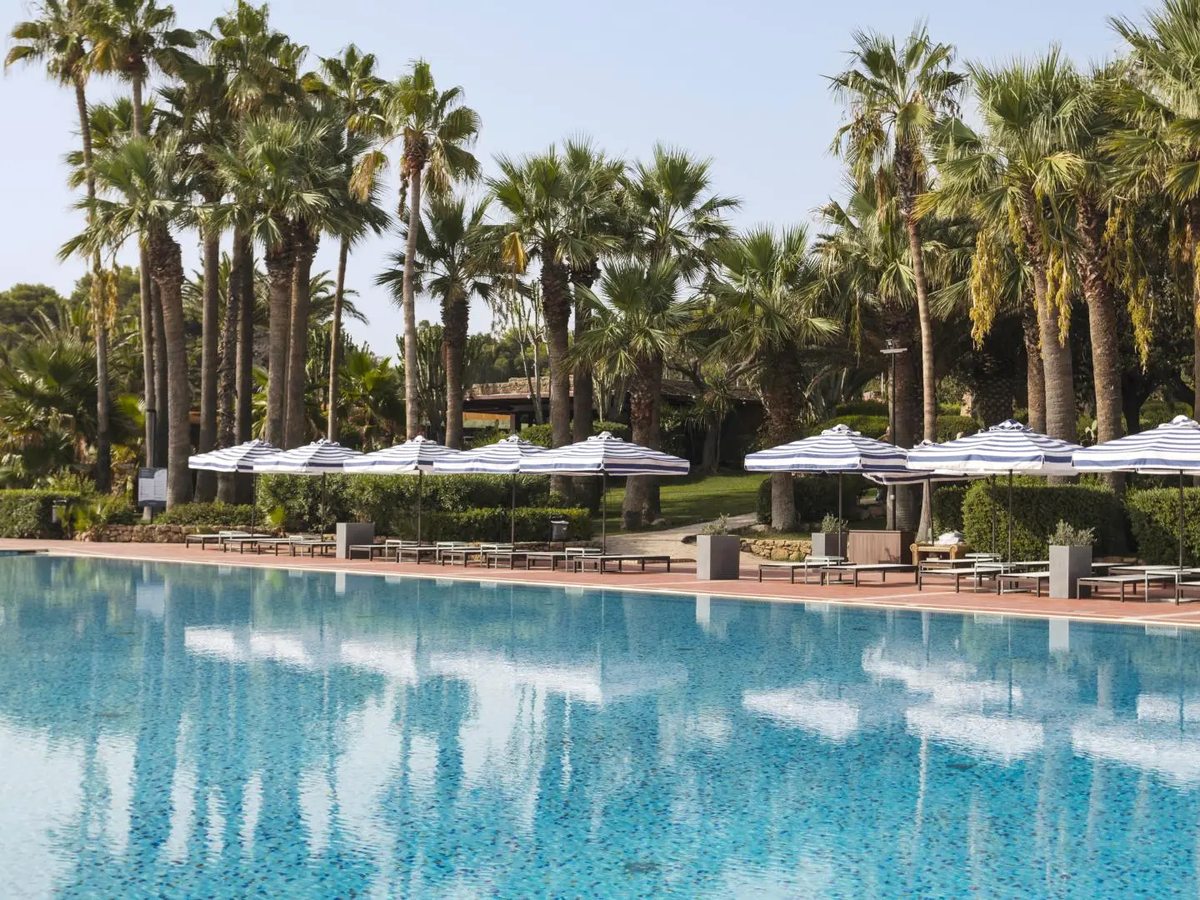 A large rectangular swimming pool with blue-and-white striped parasols and sun loungers along a terracotta-tiled surround, backed by tall palms and clipped hedges.