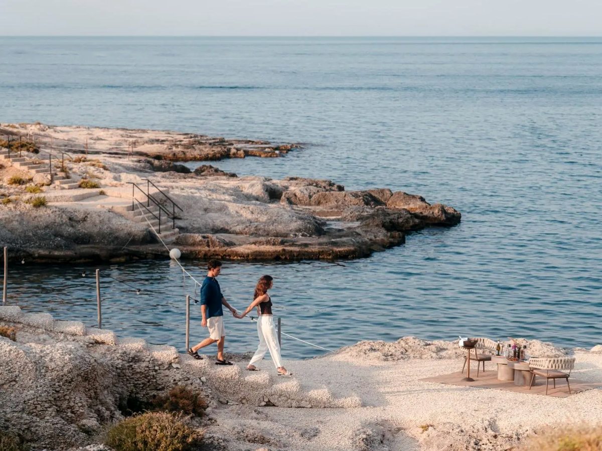A couple walking hand in hand across pale flat rocks beside a calm blue inlet, with steps cut into the rock and a low table and chairs set out on the shore.
