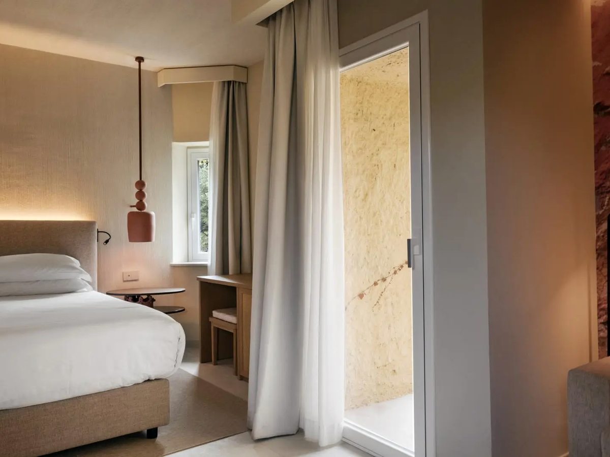 A double room with a padded headboard, terracotta pendant light and small writing desk, with a rough reddish stone wall to one side and a glazed door onto a sunlit stone-walled courtyard.