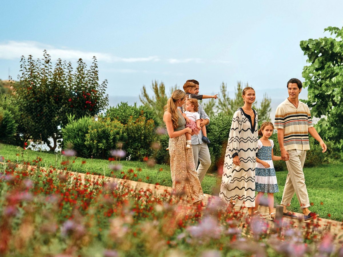 A family walking along a garden path between flowering borders and mown lawns, with mature trees and the sea beyond.