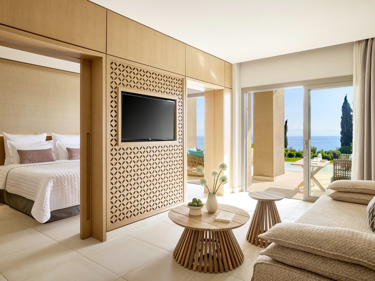 An open living area with a sofa and low round tables, the bedroom through a wide doorway, and sliding doors onto a terrace with a private pool and the sea beyond.