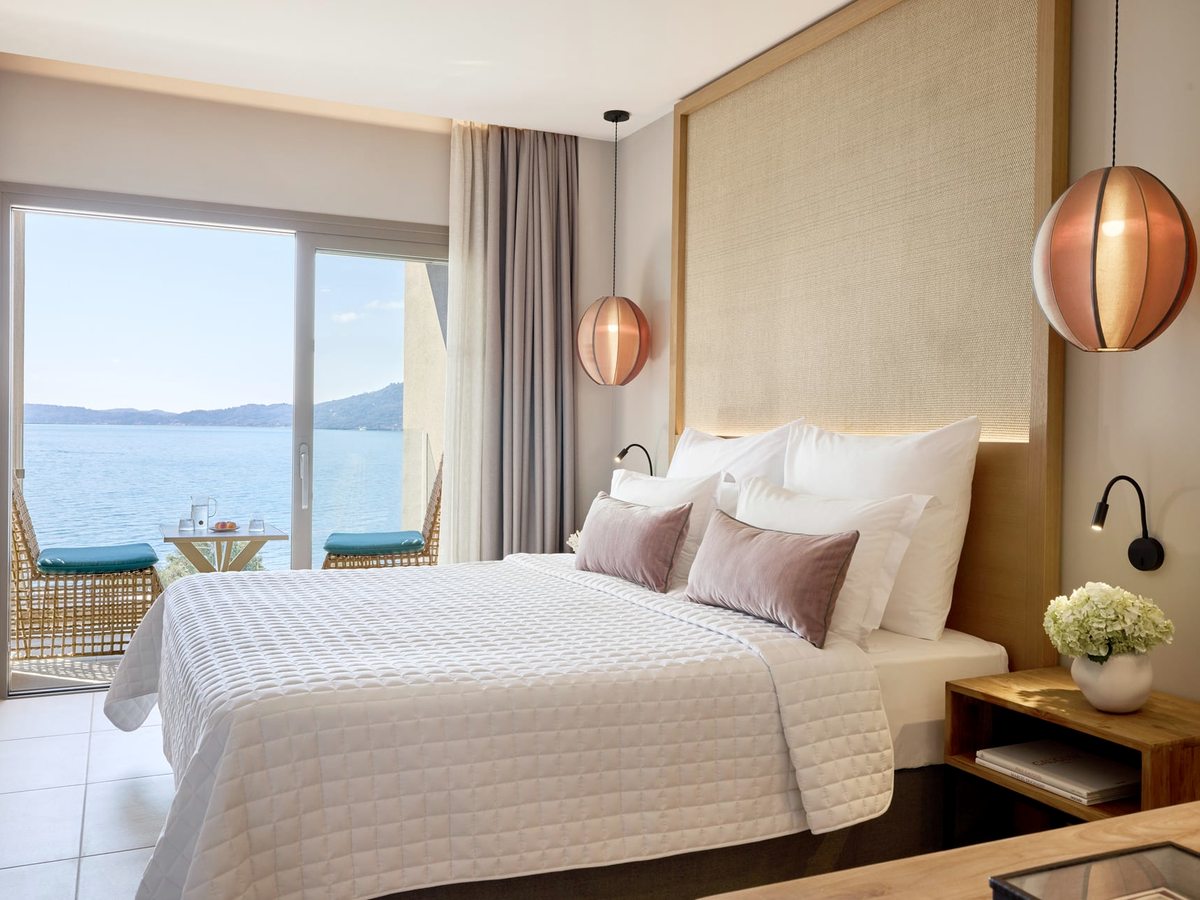 A double bed with a woven headboard and copper pendant lights, and glass doors onto a balcony with turquoise-cushioned chairs above the bay.