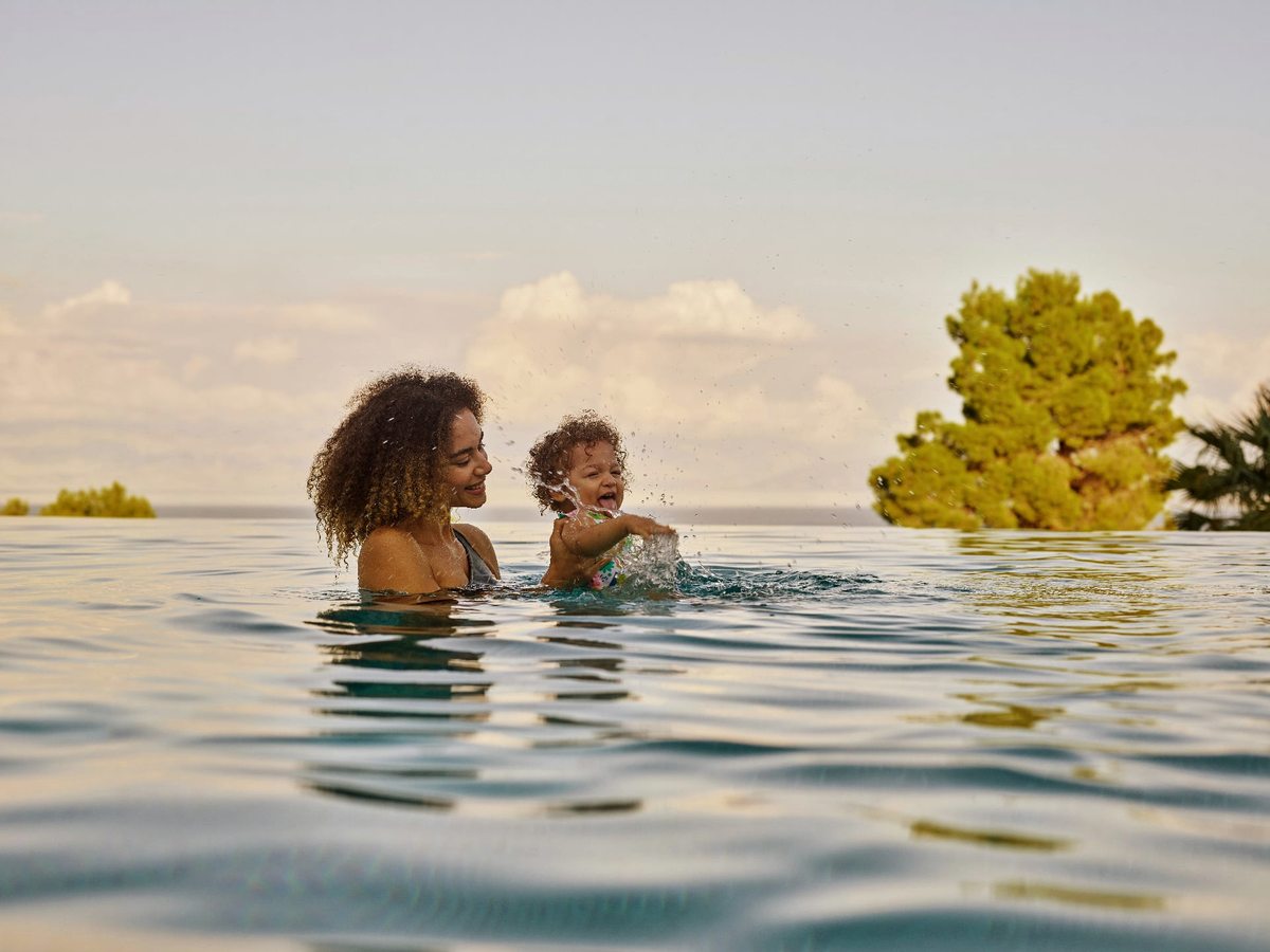 A woman holding a laughing toddler in the water at the edge of an infinity pool, with pine trees and the sea behind.