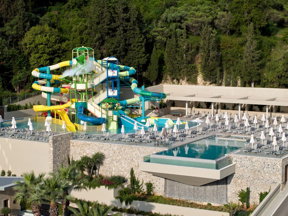 A tall cluster of yellow, blue and green water slides ending in a shallow pool, with rows of loungers and parasols on the terrace below and wooded hillside behind.