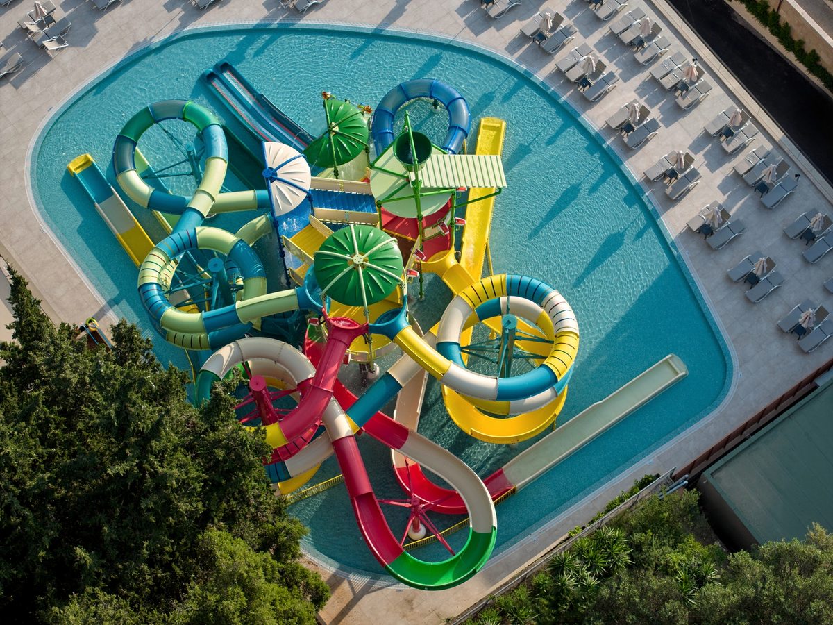 Looping red, yellow, blue and green slides set in a shallow turquoise pool, with sun loungers lined up along the paved surround.