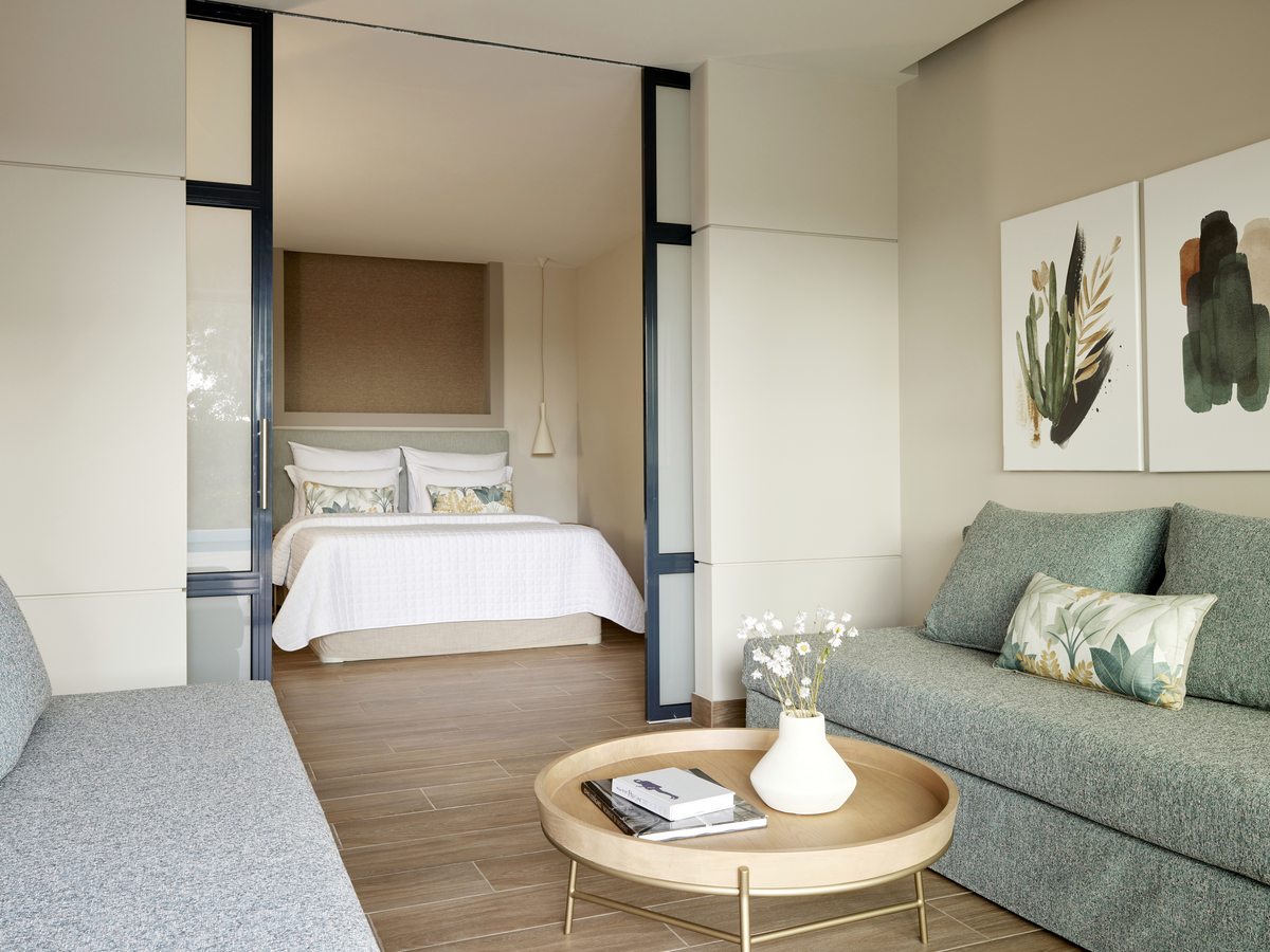A sitting room with a green sofa and a round timber table, the double bedroom visible through open sliding partitions.
