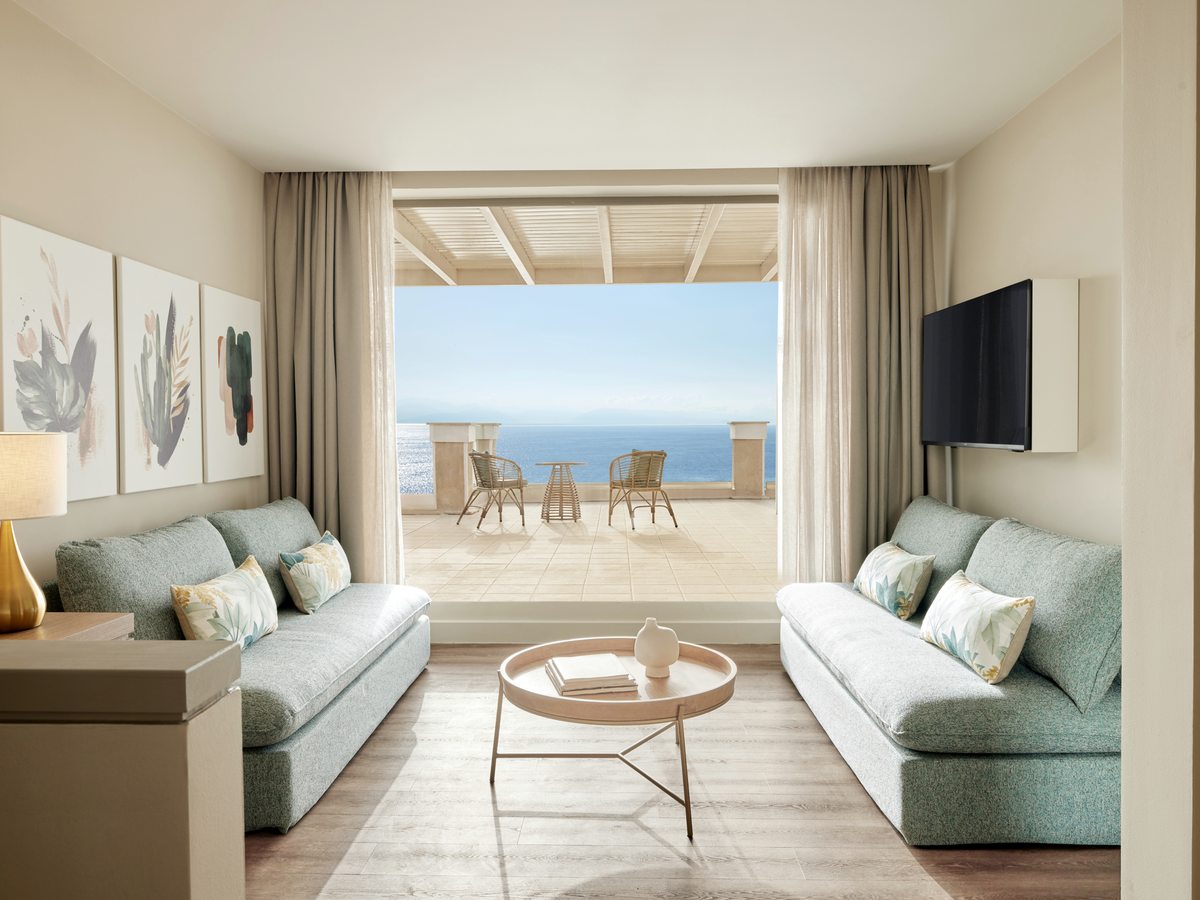 A separate sitting room with two pale green sofas either side of a low round table, opening through wide doors onto a shaded terrace above the sea.