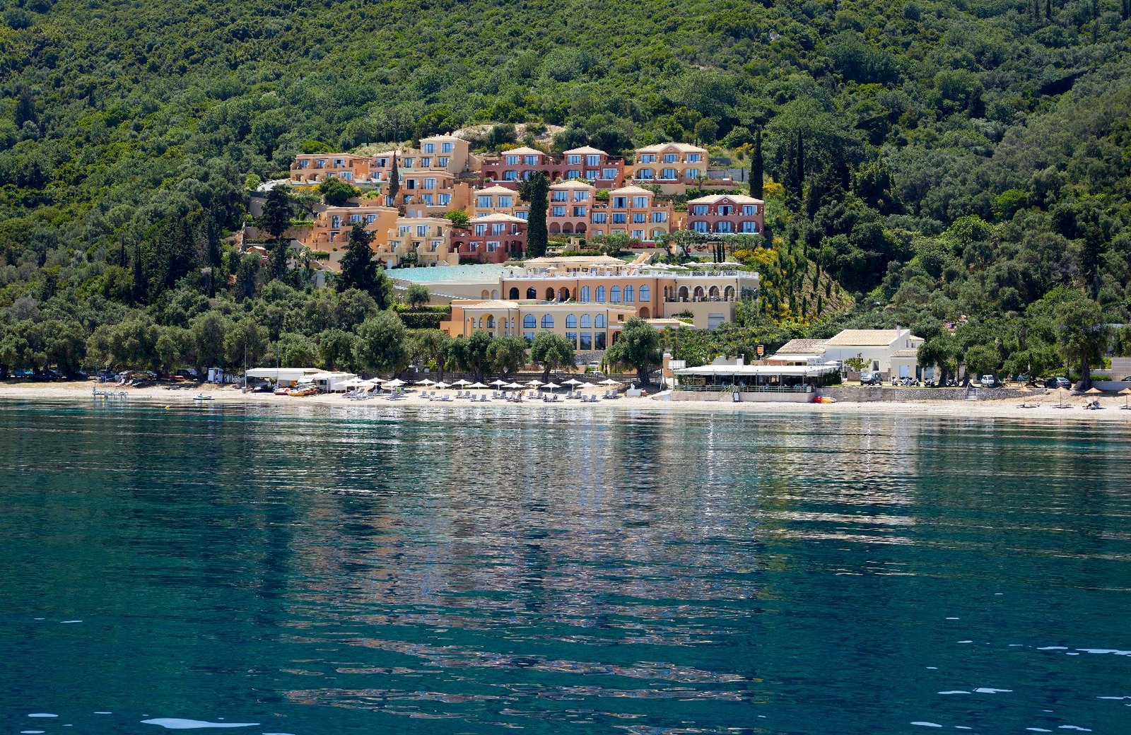 Apricot and terracotta buildings terraced up a wooded hillside above a beach lined with white parasols and loungers, with clear turquoise water in the foreground.