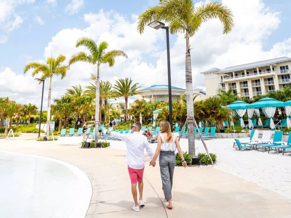 A curved lagoon-style pool with a pale sandy edge, an adult and child walking beside it, turquoise loungers, parasols and cabanas, resort buildings behind.