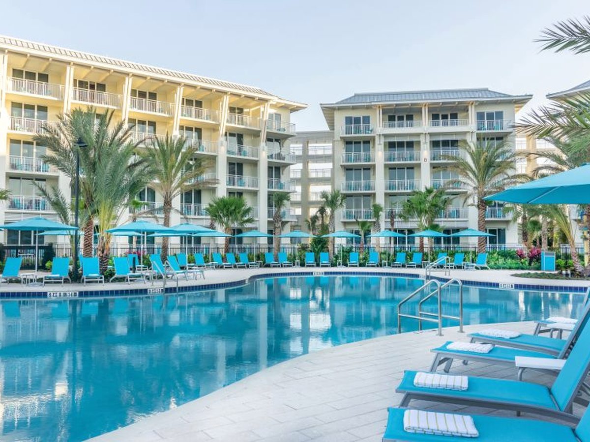 A large hotel pool with entry handrails, tiled deck, turquoise loungers and parasols, and multi-storey guest-room buildings with balconies behind.
