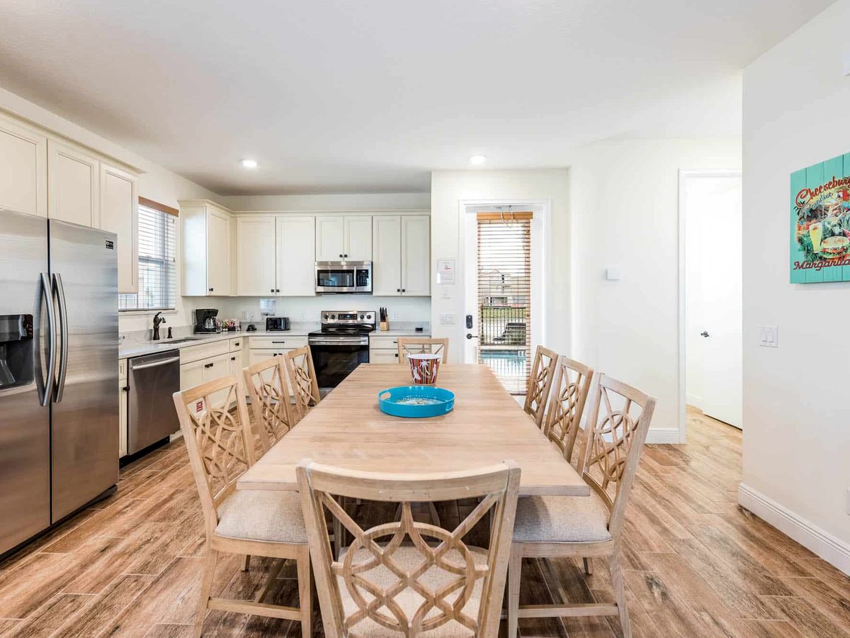 A cottage kitchen and dining area with white cabinets, a stainless-steel fridge, oven and hob, and a long wooden dining table with woven chairs.