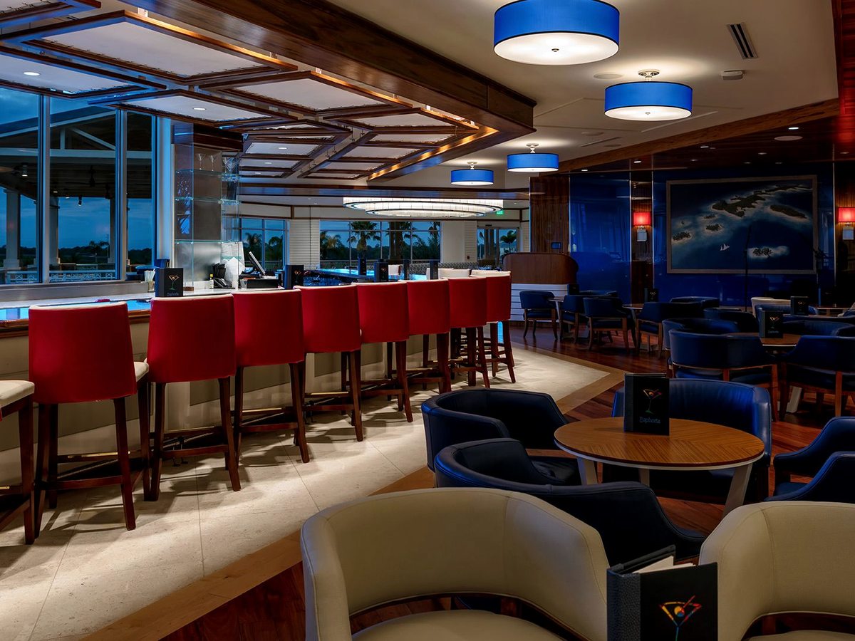 An indoor bar with a long counter, red stools, blue pendant lights, dark wood, lounge armchairs and glass walls onto the grounds.