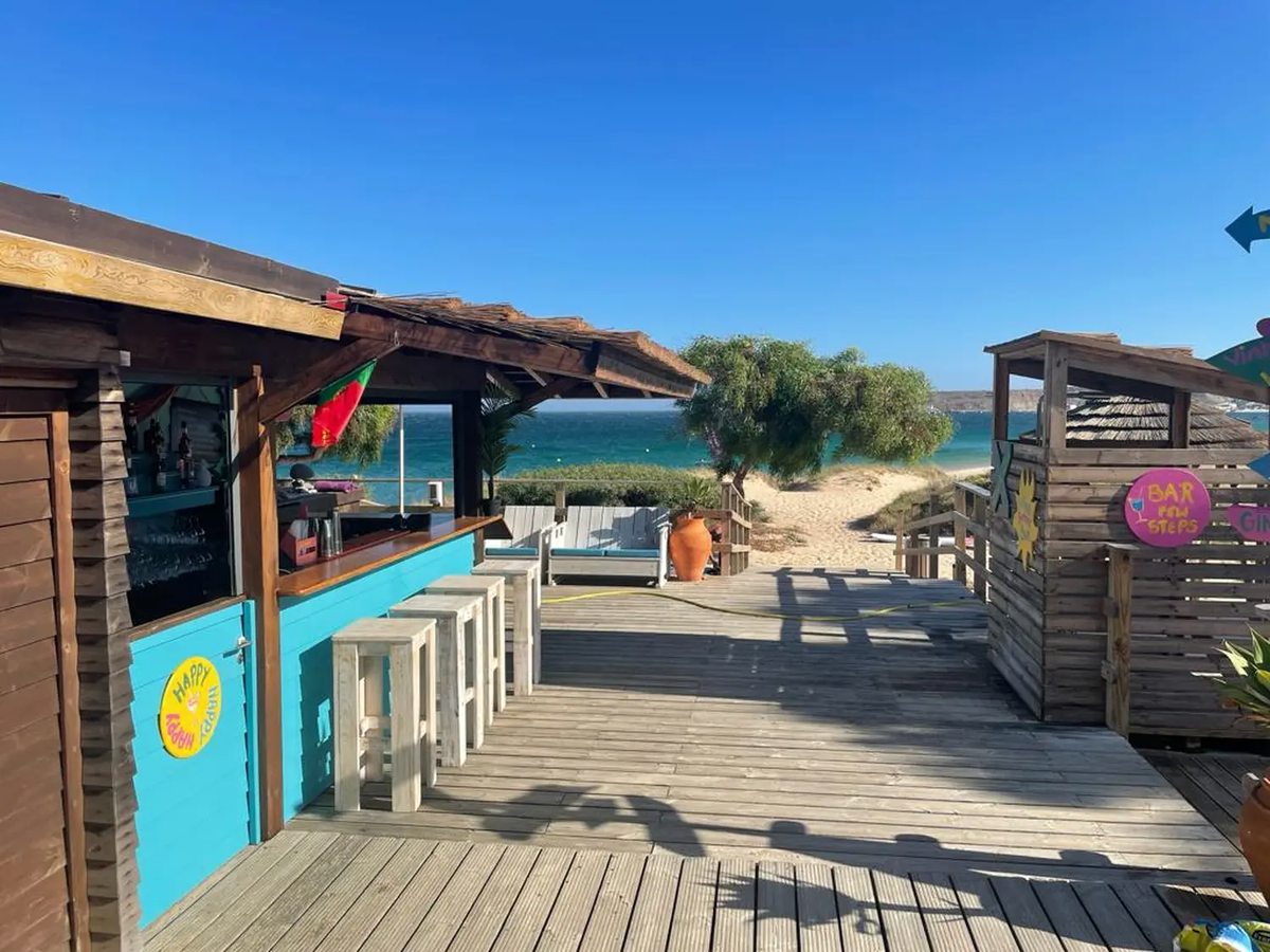 A small wooden beach bar with a turquoise counter and stools on a timber deck, painted signs, a thatched shelter and the sea beyond.