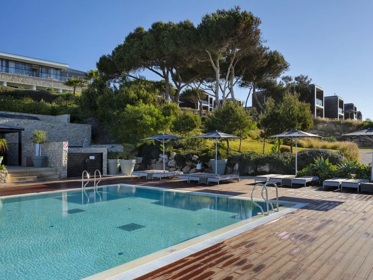 A pool on a wide timber deck with loungers and parasols, umbrella pines and the resort's terraced buildings rising on the slope behind.