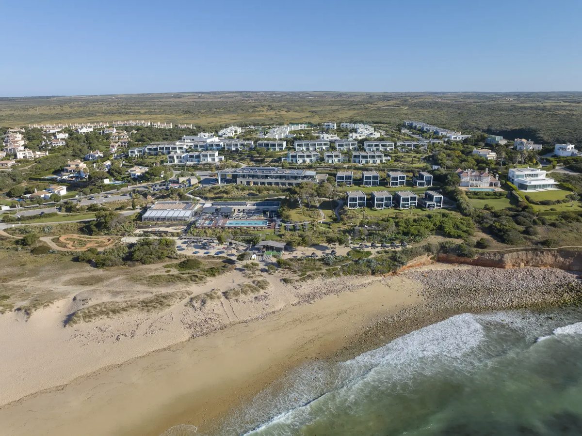 Aerial view of the resort's white flat-roofed buildings, pool terrace and gardens set behind dunes above a long sandy beach with breaking surf.