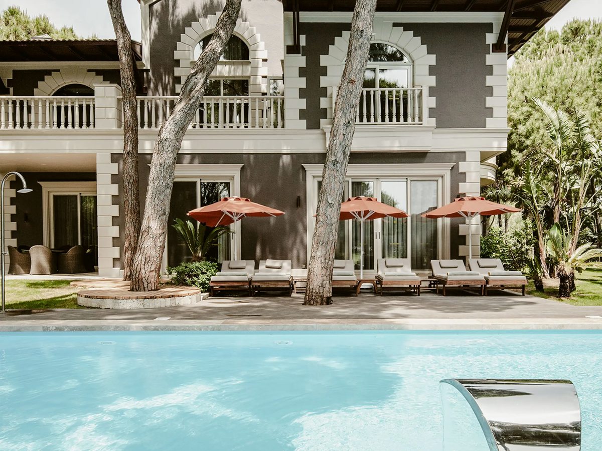 A two-storey villa with arched windows and balconies, sun loungers and coral parasols on the terrace and a private pool in front, pines overhead.