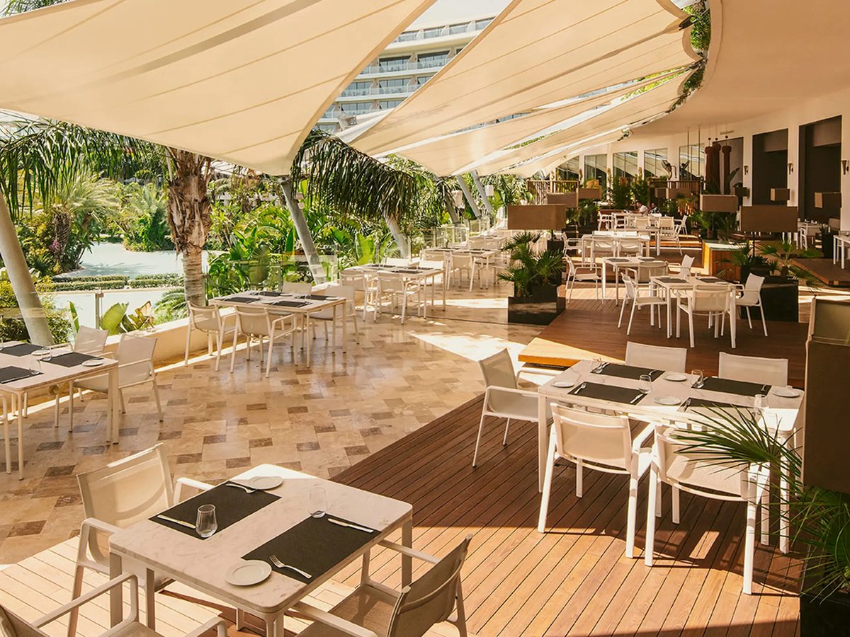 An open-air restaurant terrace under white shade sails, with white tables and chairs on timber and stone decking and palms and a pool beyond.