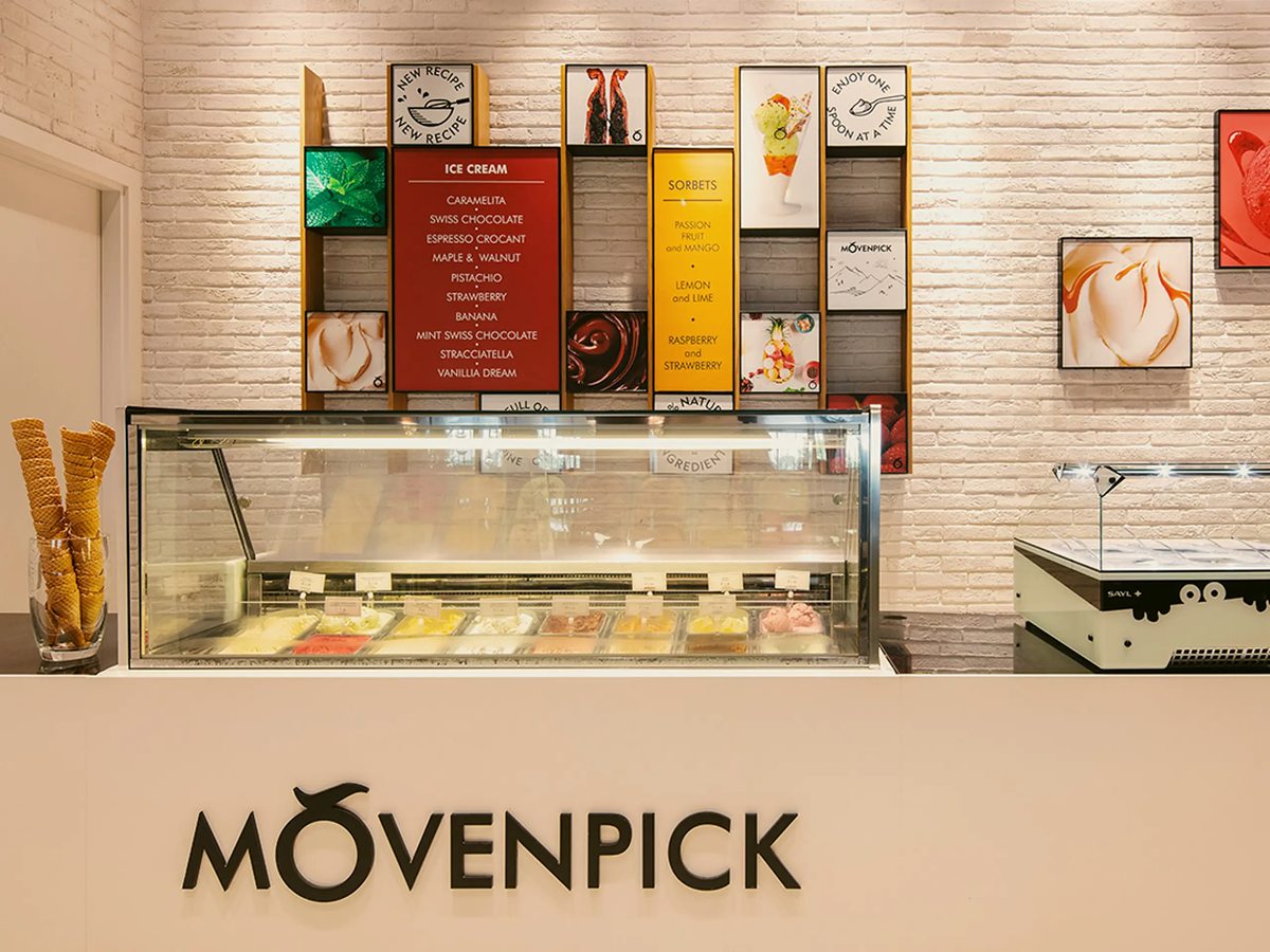A Mövenpick ice cream counter with a chilled display of tubs, waffle cones in a jar and flavour menu boards on a white brick wall.
