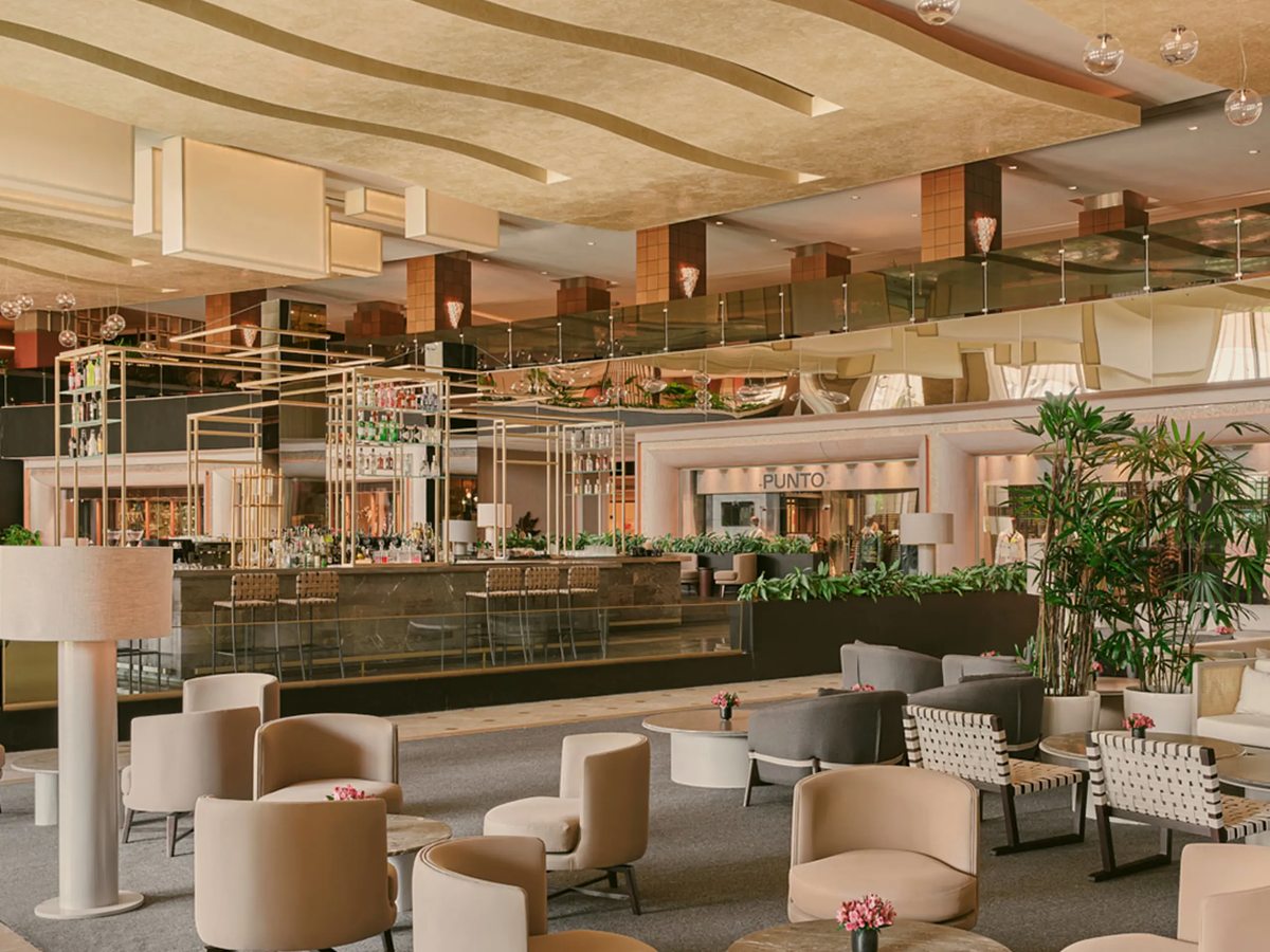 A wide lounge with a long bar, cream tub armchairs and low tables, tall planters and a mirrored ceiling.
