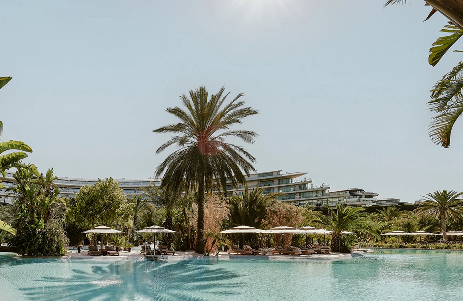 A wide turquoise pool edged with white sand and palms at Maxx Royal Belek, sun loungers and parasols along it and the curved main building behind.