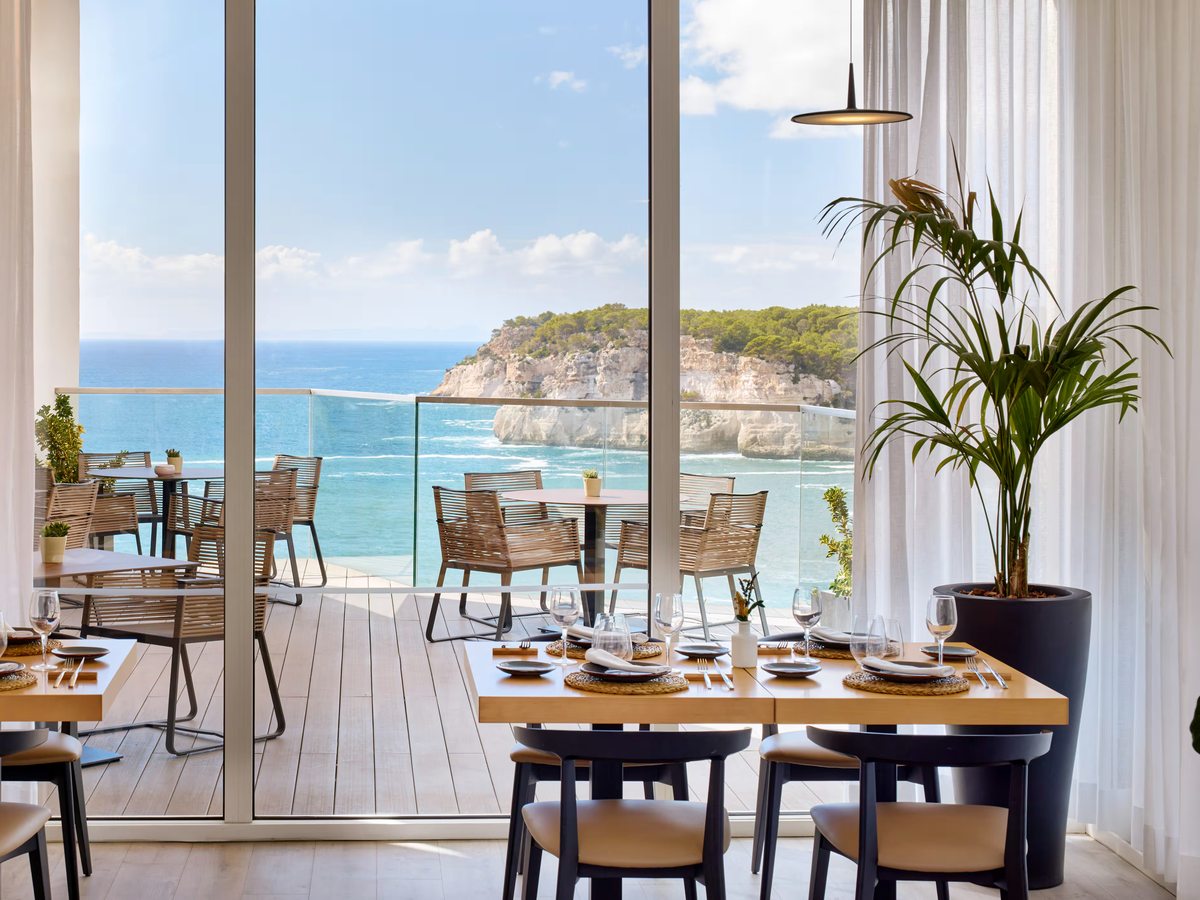 Dining room with laid tables and pale chairs behind full-height glass doors, which open onto a terrace above the sea.
