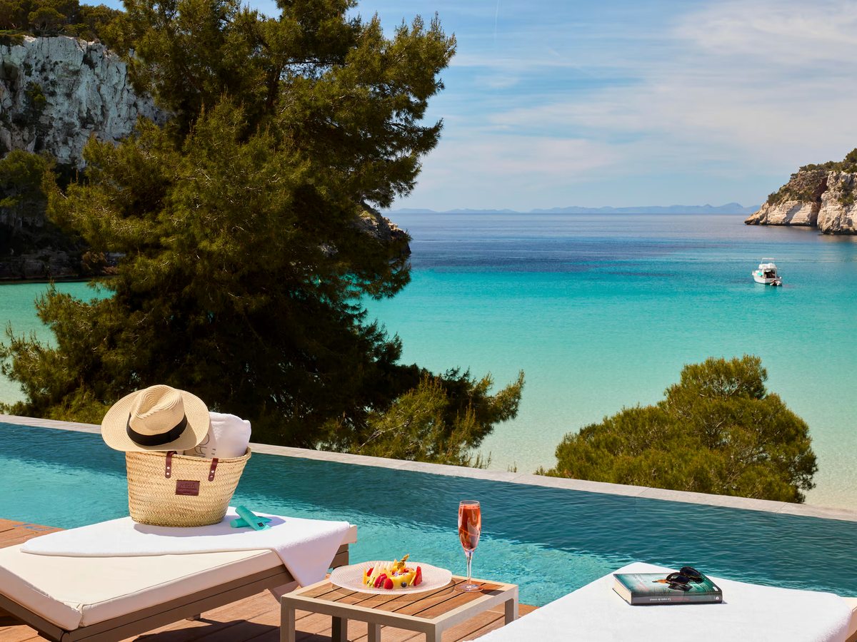 Infinity pool edge with sun loungers, a straw hat and a low table, with pines, limestone cliffs and the turquoise water of the cove behind.