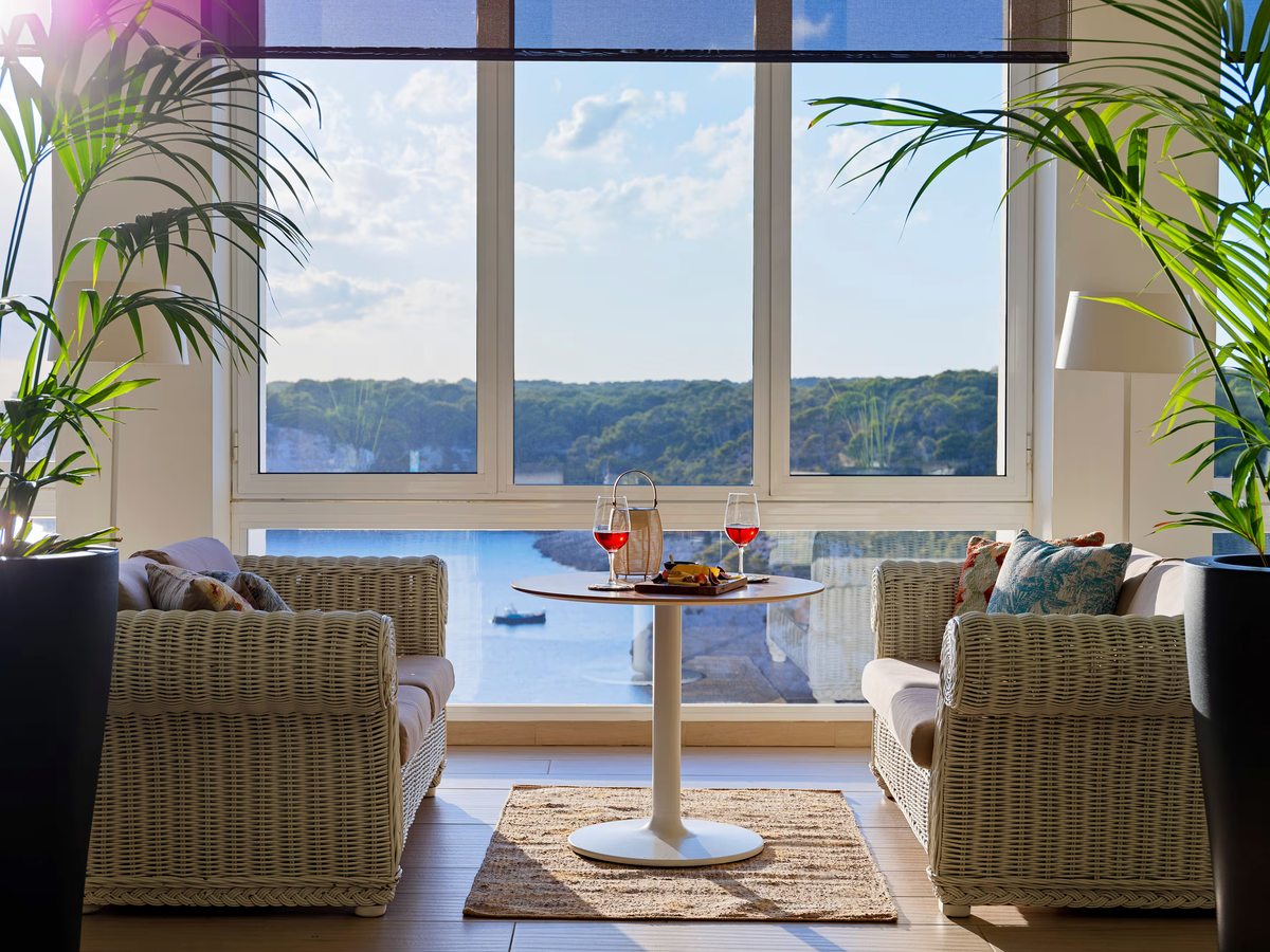 Two wicker armchairs either side of a small round table set with drinks, framed by potted palms in front of full-height windows over a wooded bay.