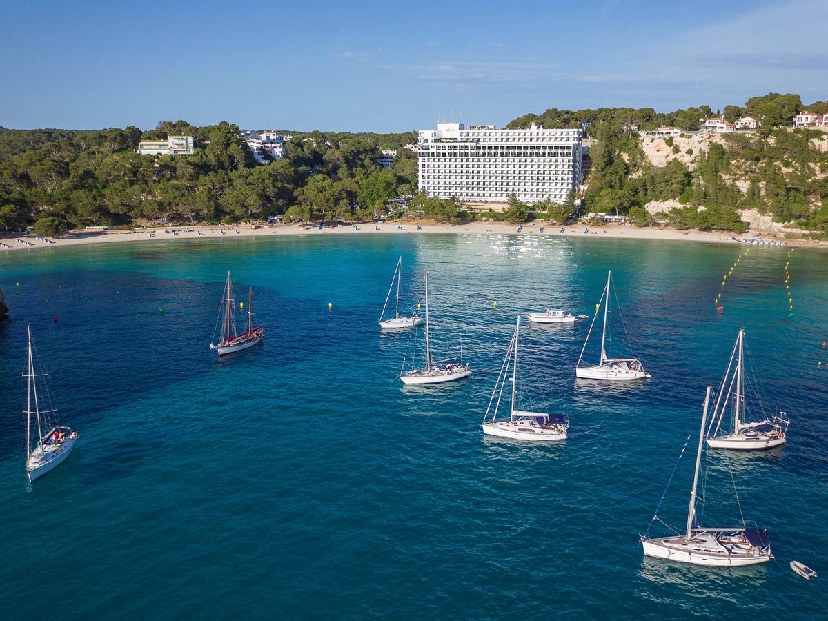 Aerial view of yachts moored in a deep blue bay, with a sandy beach and a long white hotel building on the pine-covered slope behind.