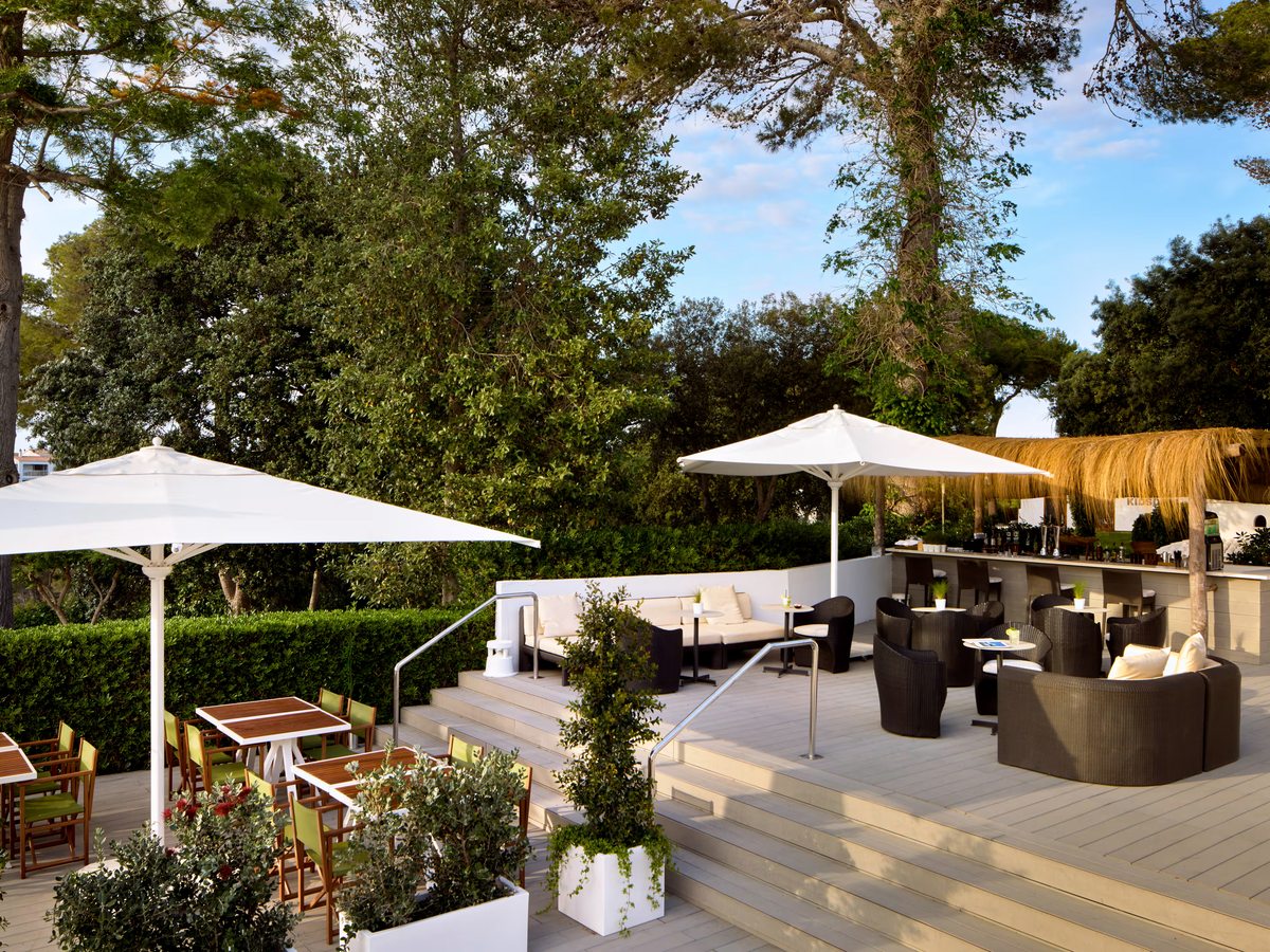 Cushioned sofas and low tables under large white parasols on a decked garden terrace, with planters and pine trees around it.
