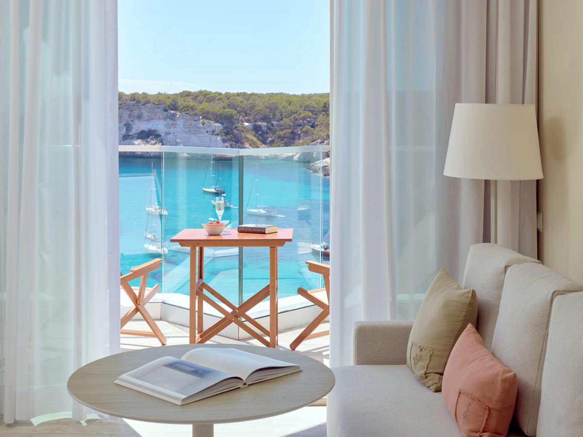 Room sitting area with a pale sofa, cushions and a round table beside open curtains, with a balcony table and chairs and the turquoise bay beyond.