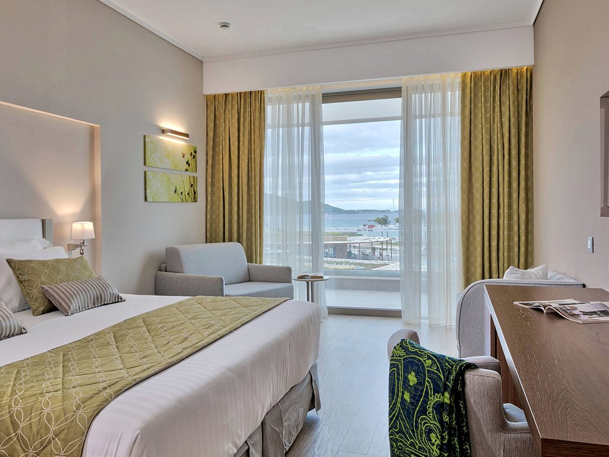 A double bed with an olive-green throw, a sofa and a desk, with sliding doors to a balcony above the coast.