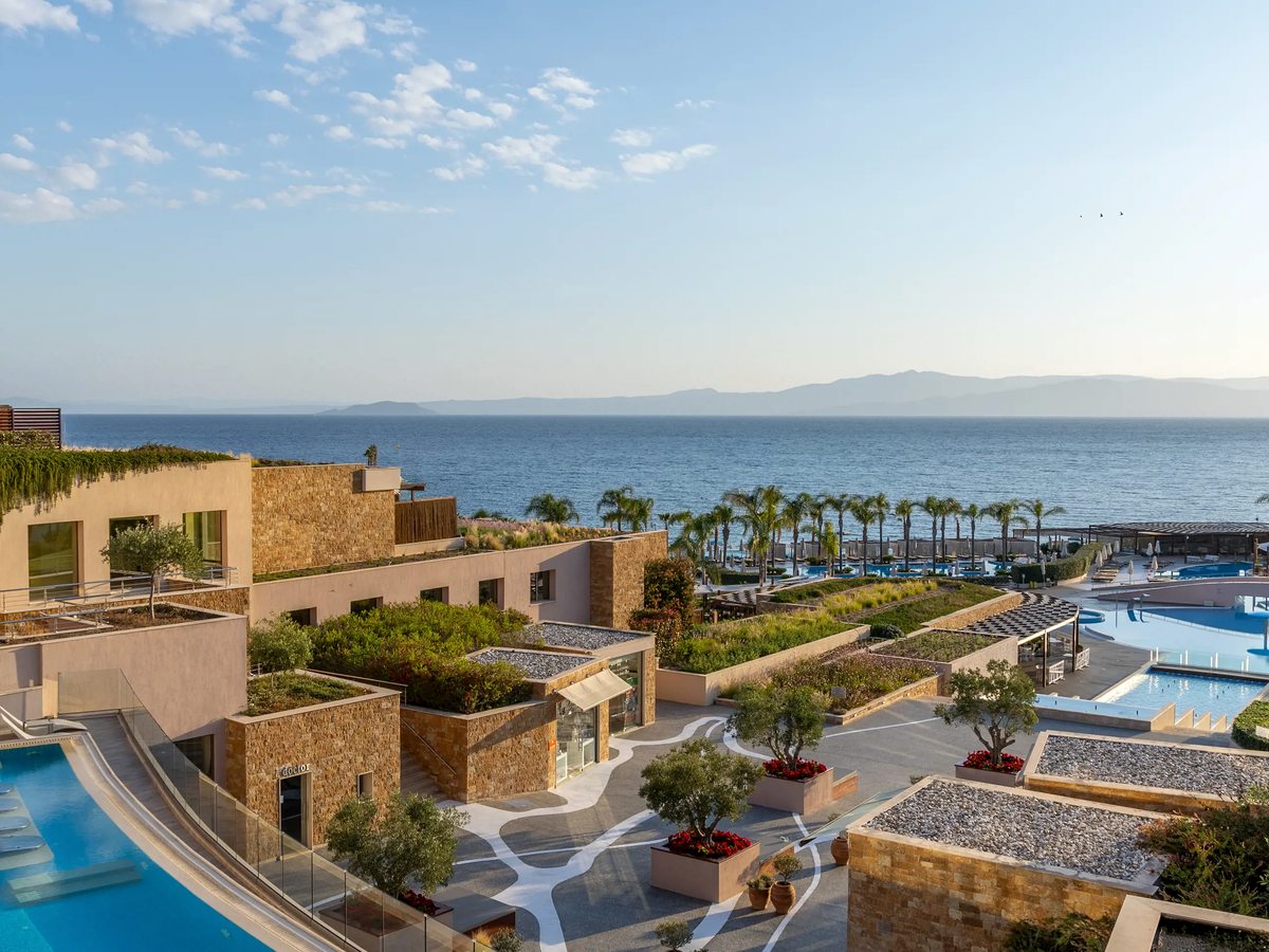 Low stone-clad guest buildings with planted green roofs and terraces, palm trees and a pool beyond, and the sea and distant hills on the horizon.