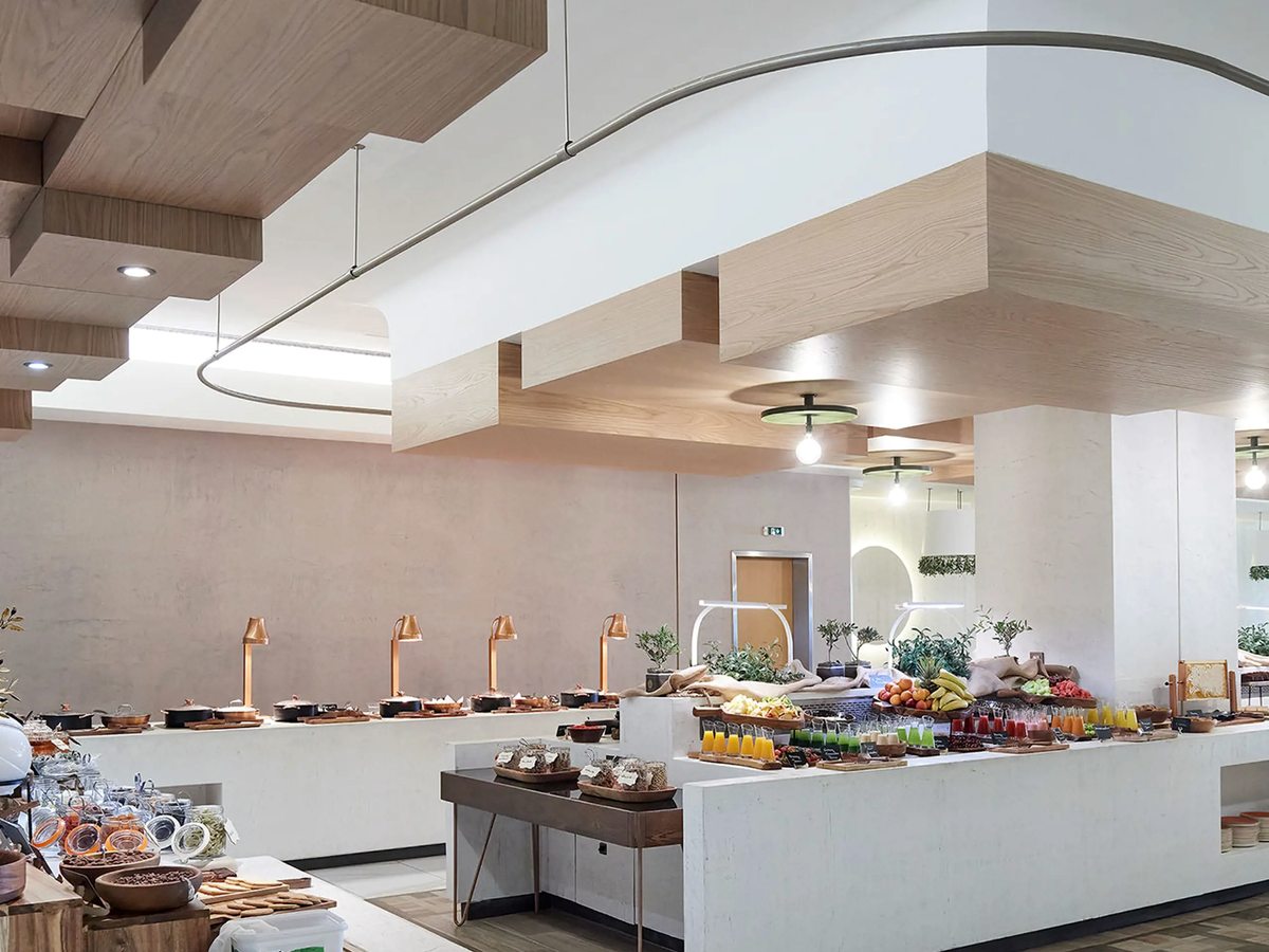 A long white buffet counter set with fruit, pastries, juices and warming lamps in a pale modern dining room.