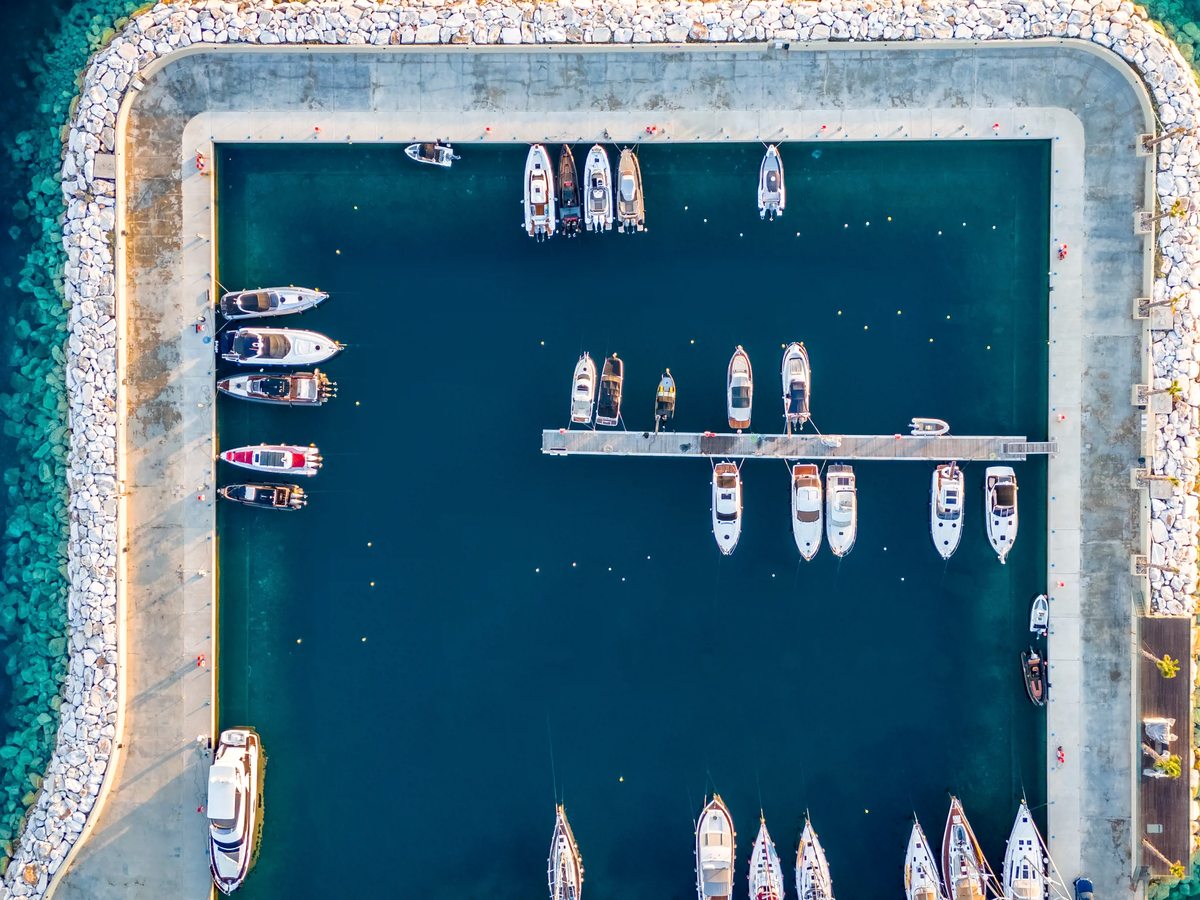The marina basin from directly above, with motor yachts and sailing boats moored along a central pontoon inside a stone breakwater.