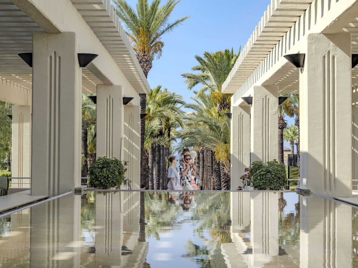 Twin white colonnades with slatted pergola roofs on either side of a still, shallow reflecting pool, with tall date palms and guests at the far end.
