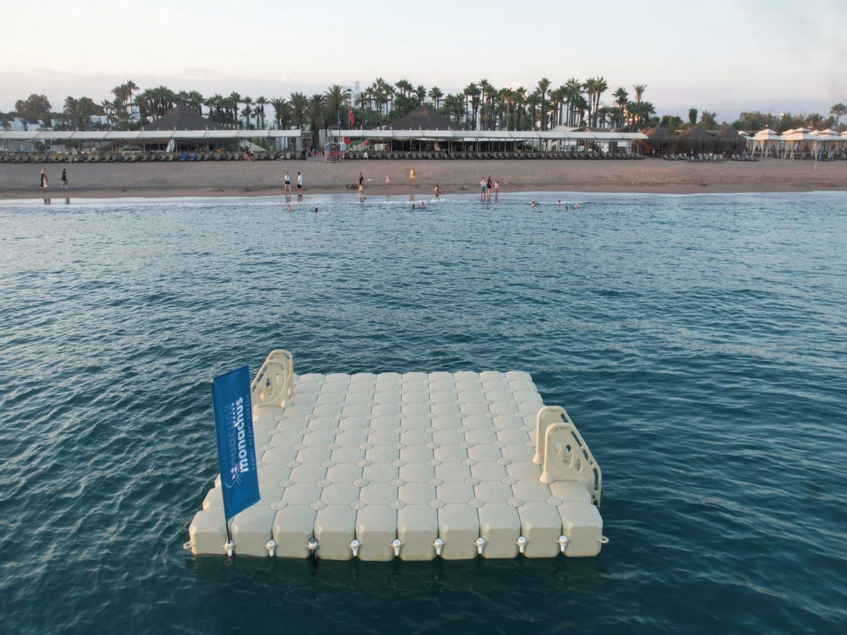 White floating swim platform anchored a short way off the beach flying a Monachus flag, with the shoreline behind.
