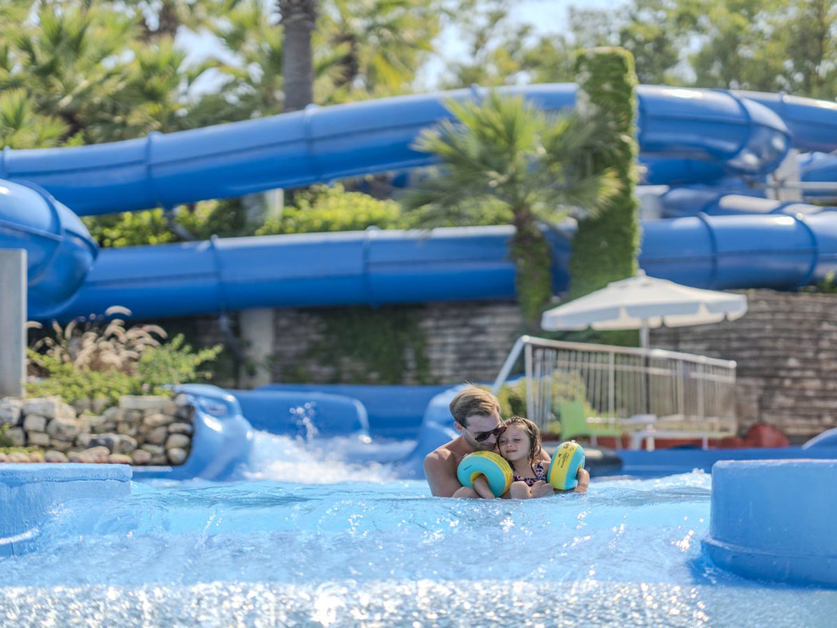 Blue water slides running down into a shallow pool where children float on rings, framed by palms and planting.