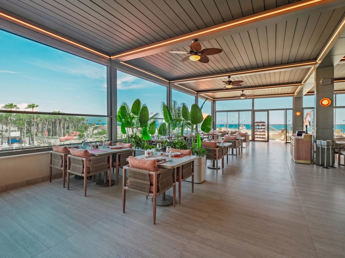 Covered rooftop restaurant terrace with cane chairs at laid tables, tall planted palms, ceiling fans and glazing open to the sea.